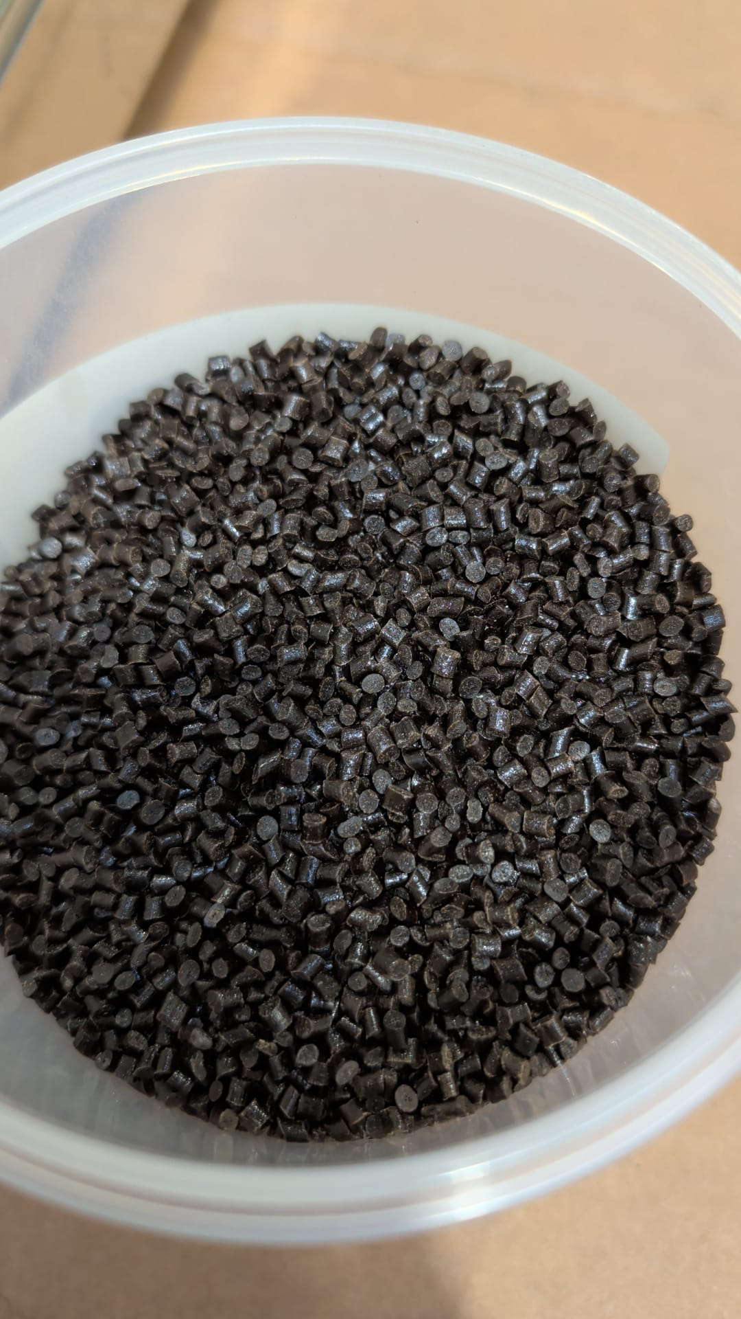 Close-up of dark cylindrical material pellets in a translucent tub on a cardboard surface