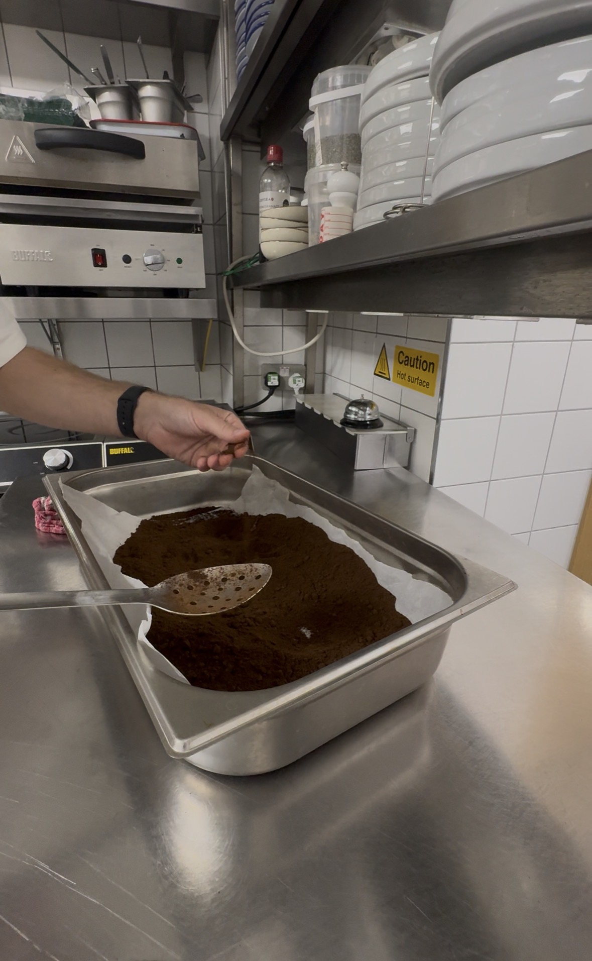 A hand adding collected coffee grounds to a parchment-lined stainless steel tray in a commercial kitchen