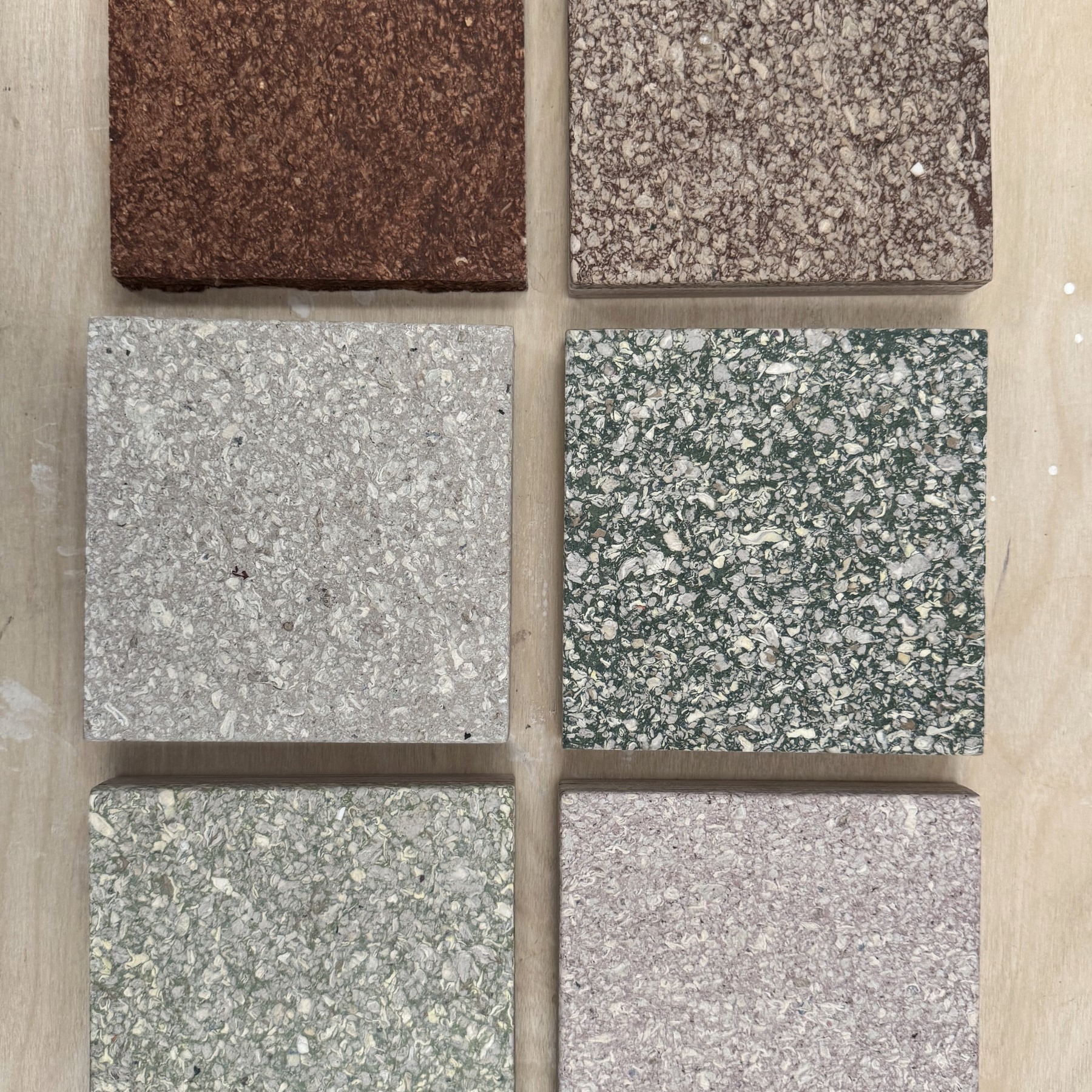 Six square MadeBy material samples in terracotta, taupe, grey, green and rose, laid on a plywood workbench