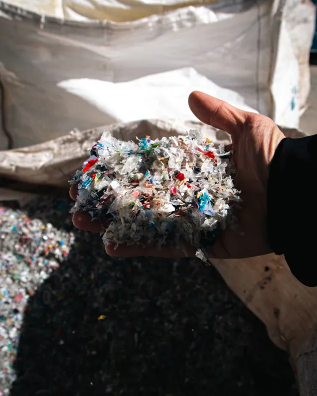 A hand holding colour-sorted plastic cuttings in front of bulk bags of recovered material