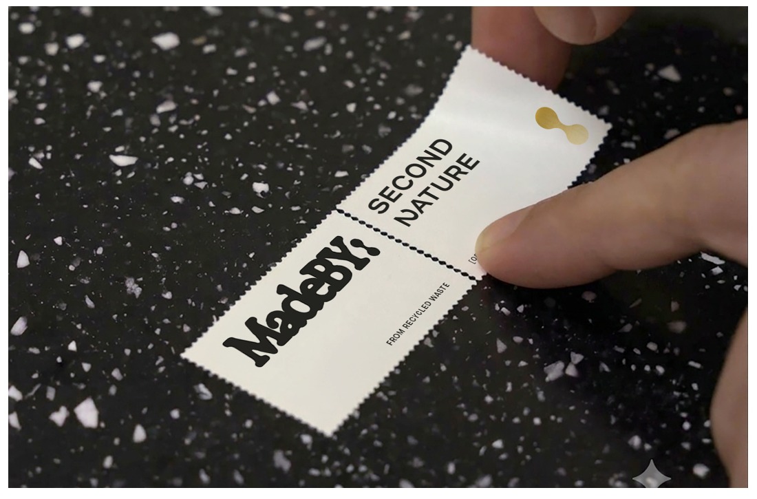 Two MadeBy labels reading MadeBy from recycled waste and Second Nature, held against the black, white-flecked recovered material they came from