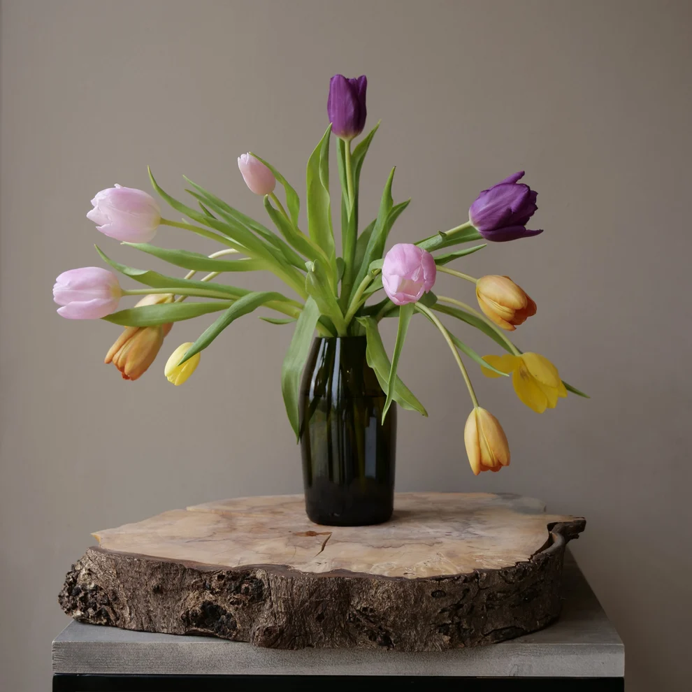 Tulips in a dark green glass vase standing on a cross-cut wood slab and a grey stone pedestal against a warm grey wall