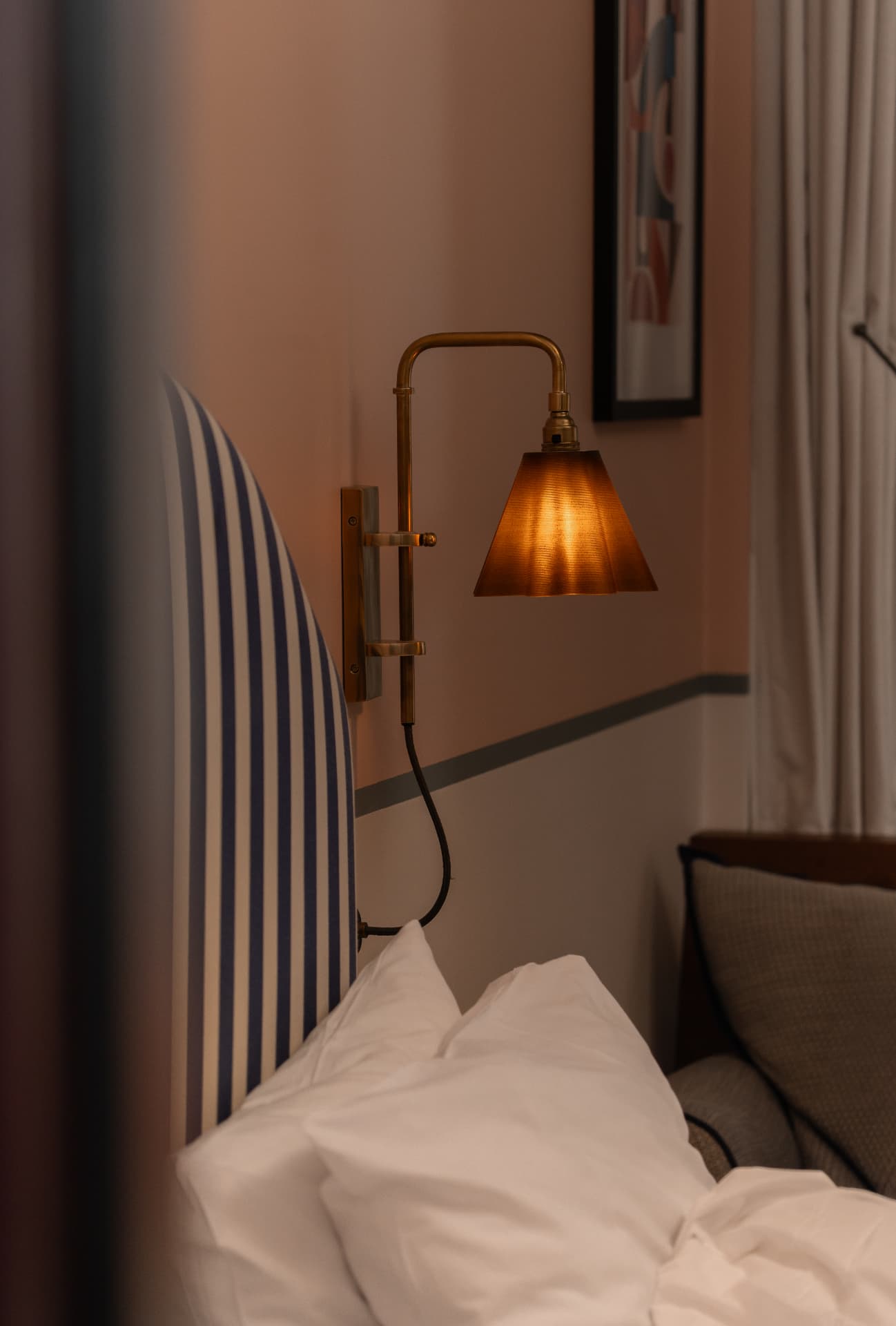 A pleated amber lamp shade glowing on a brass wall arm beside a navy-striped headboard in a hotel bedroom