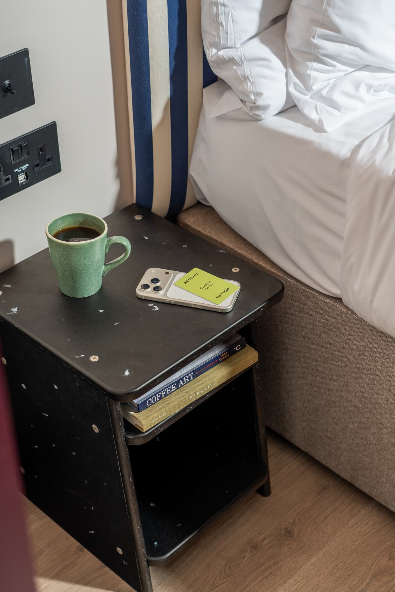 A dark recycled-plastic bedside table beside a hotel bed, holding a mug of coffee, a Second Nature keycard and two books