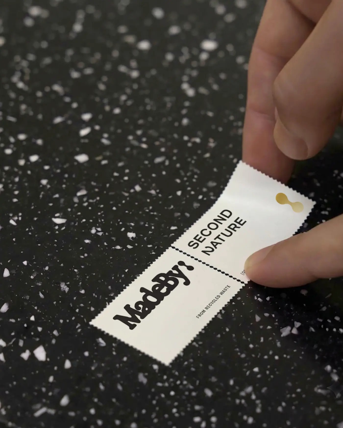 A hand holding a MadeBy material label reading From recycled waste against the black, white-flecked recovered material it came from