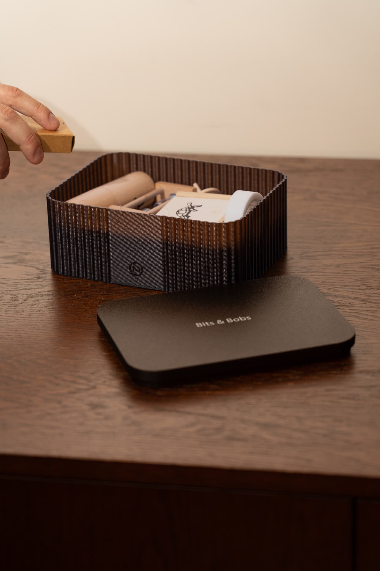 A fluted brown desktop storage box with a matching lid reading Bits & Bobs, holding wooden desk items on a dark wooden table